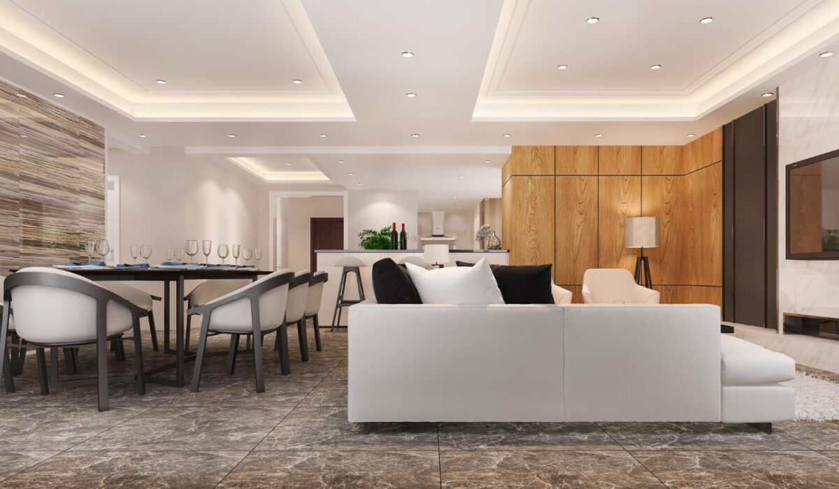Open-plan living and dining area with a white sectional sofa, black and white cushions, a long dining table, marble flooring, and a wood-paneled wall in the background.