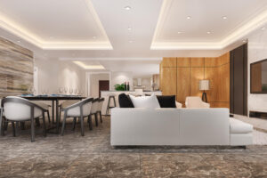 Open-plan living and dining area with a white sectional sofa, black and white cushions, a long dining table, marble flooring, and a wood-paneled wall in the background.