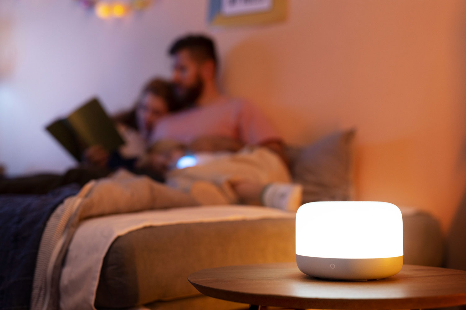 Dimly lit bedroom with a glowing bedside lamp on a wooden table; blurred couple reading in bed in the background.