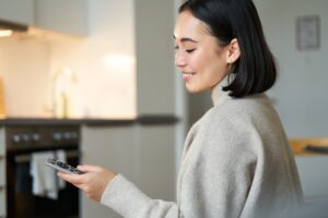 Woman in a beige sweater holds a remote and smiles, standing in a kitchen.